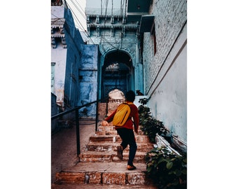 May include: A young person wearing a red shirt and a yellow backpack walks up a set of stone steps in a narrow alleyway. The walls of the alleyway are painted blue and white.