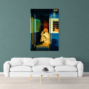 May include: A man sits on the floor of a train car, wearing a white robe and a pink turban. The train car is blue and has a window with a yellow frame. The man is looking down at his hands.