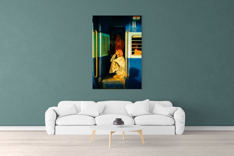 May include: A man sits on the floor of a train car, wearing a white robe and a pink turban. The train car is blue and has a window with a yellow frame. The man is looking down at his hands.