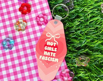 Könnte beinhalten: Ein rosa Schlüsselanhänger im Motel-Stil mit dem Text "HOT GIRLS HATE FASCISM" in einem roten Herz. Der Schlüsselanhänger liegt auf einer rosa-weiß karierten Oberfläche, umgeben von bunten blütenförmigen Perlen.