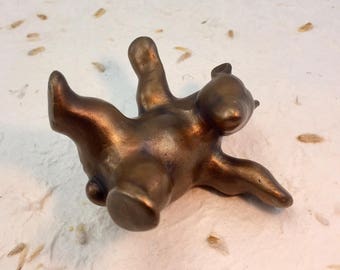 May include: A small, solid bronze-colored figurine of a bear in a reclining position. The bear is depicted lying on its back with its limbs extended upward and outward, featuring a smooth, metallic finish with subtle dark shading in the crevices.
