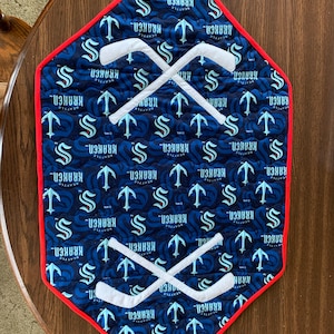 May include: A diamond-shaped blue and white fabric table runner with a red border. The fabric features a repeating pattern of the Seattle Kraken hockey team logo, which includes a stylized 'S' and an anchor. The runner also features two white hockey sticks crossed in the center.
