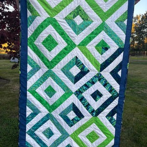 May include: A handmade quilt featuring a geometric pattern of green and white squares and diamonds. The quilt has a dark blue border and is held up against a background of green grass and trees. The quilt is approximately 48 inches long and 36 inches wide.