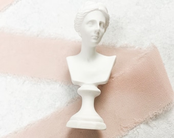 May include: White plaster bust of a woman with a simple, elegant design. The bust is on a white pedestal and is set against a background of pink fabric.