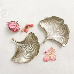 May include: Two silver ginkgo leaf-shaped trays with pink flower petals on a white background.