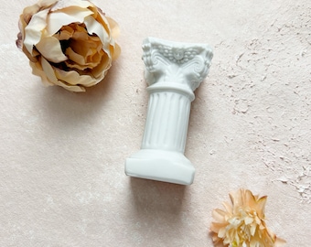 May include: A white ceramic column with decorative details sits on a light pink textured surface with two beige fabric flowers.