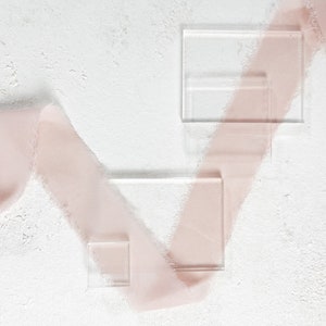 May include: Clear acrylic rectangles of varying sizes are arranged on a textured white surface, with a soft pink ribbon weaving across the scene. The composition creates a minimalist, elegant aesthetic, suitable for product display or artistic presentation.