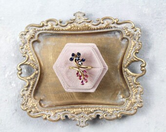 May include: A gold ring with a delicate, twisting design set with blue and pink gemstones. The ring is displayed in a pink velvet ring box on a gold tray.
