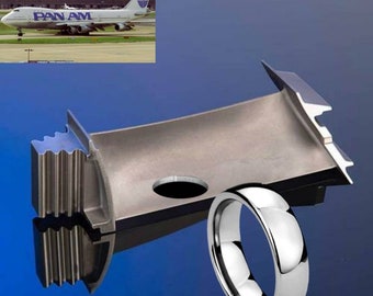 May include: A silver metal airplane engine part with a round hole and a silver metal ring next to it. The engine part is on a blue surface. The image also includes a photo of a Pan Am airplane in the background.