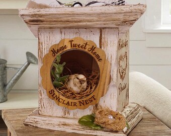 May include: A distressed white and pink wooden birdhouse with the words "Home Sweet Home" and "Sinclair Nest" on the front. The birdhouse has a round opening with a nest inside. A small decorative nest sits on a wooden table.