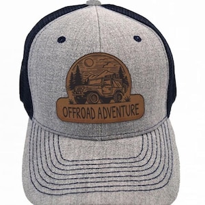 Puede incluir: Gorra de camionero gris y negro con un parche de cuero marrón que dice "Offroad Adventure". El parche presenta una silueta de un jeep conduciendo por un paisaje montañoso.