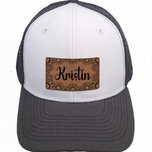 May include: White and grey trucker hat with a brown leather patch that says "Kristin" in black script. The patch is decorated with a floral design.