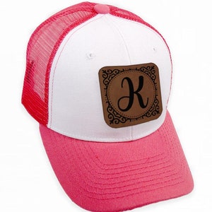 Peut inclure: Casquette de camionneur blanche et rose avec un patch en cuir marron orné d'une lettre K noire dans un cadre décoratif.