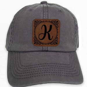 Op de afbeelding: Een donkergrijze baseballpet met een bruine leren patch met een decoratieve rand en de letter "K" in een cursief schrift.