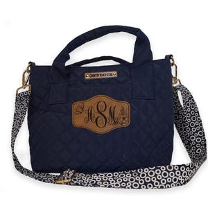 Op de afbeelding: Marineblau gewatteerde tote bag met een bruin leren label met de initialen "ASM" en bloemmotieven. De tas heeft een patroonband en goudkleurige hardware. De tekst "COUNTRY TOUCH STORE O" is zichtbaar.