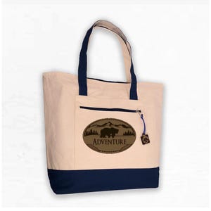 May include: A beige canvas tote bag with a navy blue bottom. The bag has a brown leather patch with a silhouette of a bear and mountains, and the text "ADVENTURE".