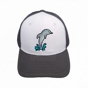 Könnte beinhalten: Eine weiße und graue Trucker-Kappe mit einem grauen Netzrücken. Die Vorderseite der Kappe zeigt ein gesticktes Bild eines grauen Delfins, der aus dem Wasser springt, mit blauen und grünen Wellen.