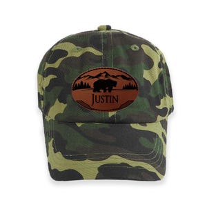 May include: Camouflage baseball cap with a brown leather patch featuring a silhouette of a bear in front of mountains and the name "Justin".