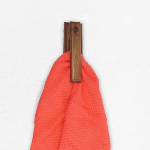 May include: A wooden towel clip with a dark brown finish. The clip is mounted on a white wall and has a coral colored towel hanging from it.
