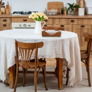 May include: A wooden tissue box holder with a white tissue box inside. The holder is on a white tablecloth on a wooden table with four wooden chairs. There are flowers in a white pitcher on the table.