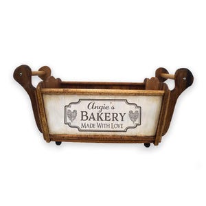 Puede incluir: Cesta de panadería de madera con forma rectangular y asas curvas. El panel frontal presenta el texto "Angie's Bakery MADE WITH LOVE" en una fuente decorativa. La cesta está hecha de madera marrón clara y oscura.