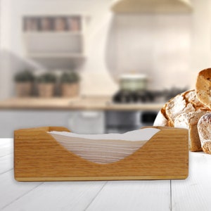 May include: A wooden napkin holder with a white napkin inside. The holder is made of light brown wood and has a curved opening for the napkins. The holder is sitting on a white wooden table.