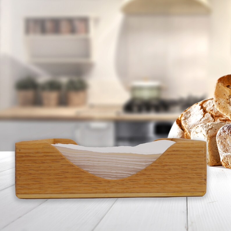 May include: A wooden napkin holder with a white napkin inside. The holder is made of light brown wood and has a curved opening for the napkins. The holder is sitting on a white wooden table.
