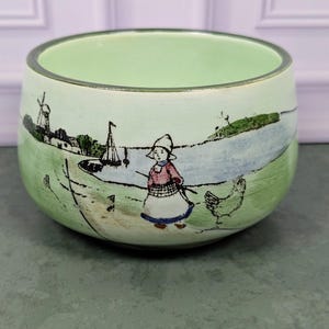 May include: A light green ceramic bowl featuring a hand-painted scene of a person in a bonnet and apron standing near a body of water with a sailboat, a windmill, and a chicken. The bowl has a smooth, rounded shape and a dark rim along the top edge.