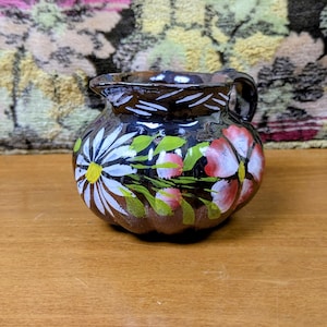 May include: A small, dark brown ceramic creamer pitcher with a glossy finish, featuring hand-painted white daisy flowers and pink blossoms with green leaves. The pitcher has a rounded, ribbed body, a small pouring spout, and a curved handle.