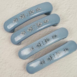 May include: Four arched cabinet handles in a smooth, glossy blue finish. Each handle features a centered, repeating silver-toned floral pattern consisting of stylized leaves and small dots, arranged in a linear design along the top surface.