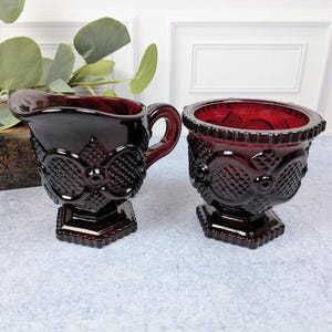 May include: A deep red glass creamer and sugar bowl set featuring a raised diamond pattern and circular motifs. Both pieces have a scalloped rim, a hexagonal base, and a rich, translucent ruby color that catches the light with a glossy finish.