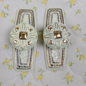 May include: Two decorative cabinet pulls. Each pull has a rectangular base with a light green finish and gold trim. A round, ornate knob with a gold square center is attached to each base. The pulls are on a white floral patterned background.