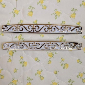 May include: Two decorative metal hair barrettes with a silver and gold filigree design. The barrettes are rectangular and feature an ornate, repeating pattern. They rest on a cream-colored floral patterned fabric.