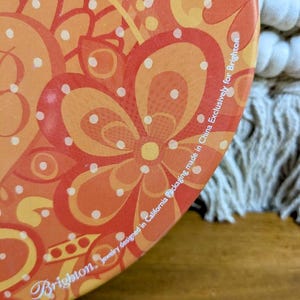 May include: Close-up of a round, orange and yellow floral patterned container. The design features large flower shapes and polka dots. The text "Brighton. Jewelry designed in California Packaging made in China Exclusively for Brighton" is printed on the side.