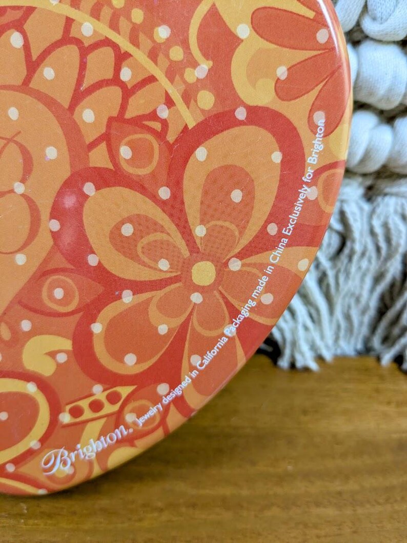 May include: Close-up of a round, orange and yellow floral patterned container. The design features large flower shapes and polka dots. The text "Brighton. Jewelry designed in California Packaging made in China Exclusively for Brighton" is printed on the side.