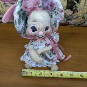 Puede incluir: Mu&ntilde;eca de conejito hecha a mano con un gorro y un vestido florales. La mu&ntilde;eca tiene una cara pintada con una nariz rosa y una peque&ntilde;a sonrisa. Las orejas de la mu&ntilde;eca son rosas y mide aproximadamente 7.6 cm de alto.