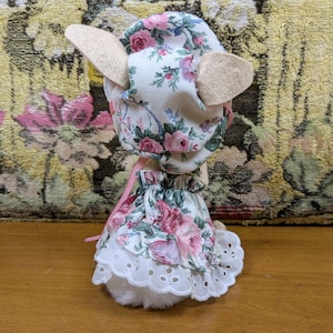 Puede incluir: Un cordero de peluche con un gorro y un vestido con estampado floral. El gorro y el vestido tienen un fondo blanco con rosas rosas y moradas y hojas verdes. El cordero tiene orejas beige y un cuerpo blanco y esponjoso.