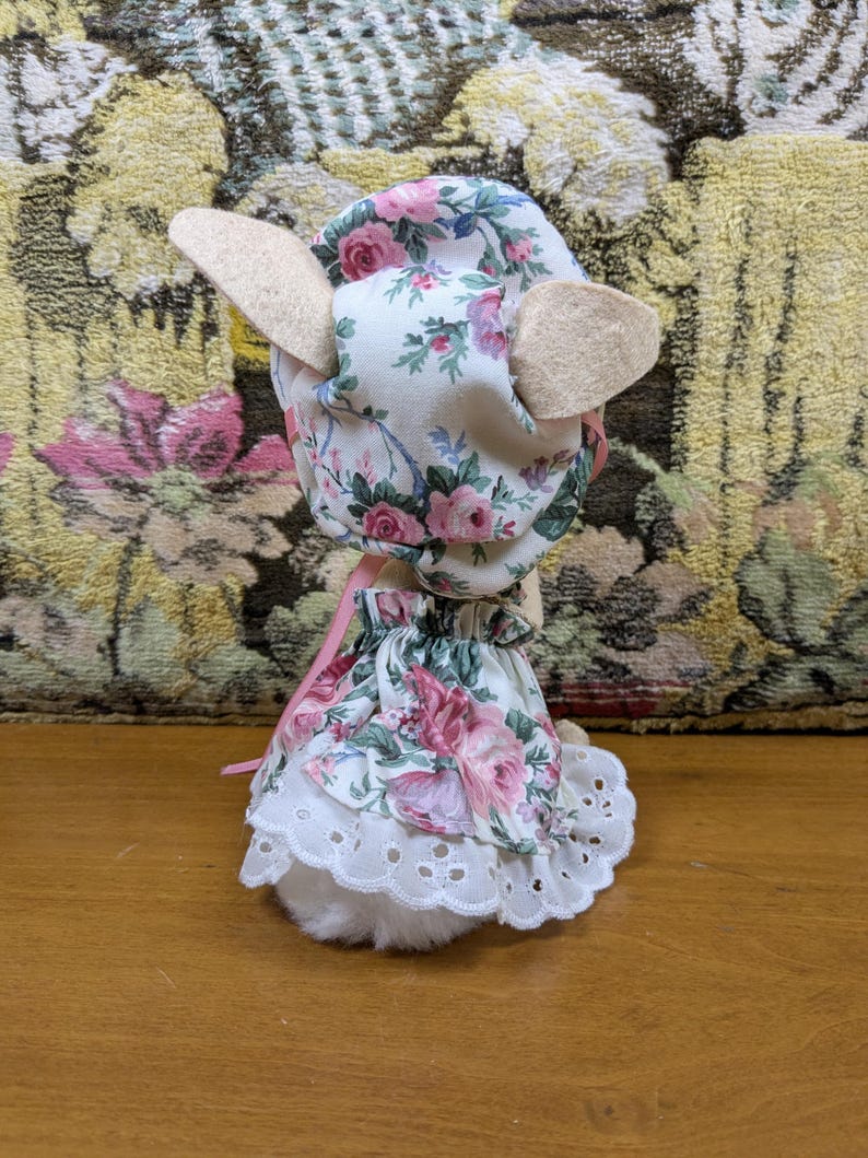 Puede incluir: Un cordero de peluche con un gorro y un vestido con estampado floral. El gorro y el vestido tienen un fondo blanco con rosas rosas y moradas y hojas verdes. El cordero tiene orejas beige y un cuerpo blanco y esponjoso.