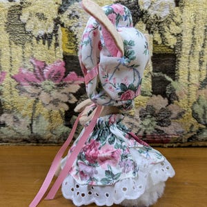 Puede incluir: Una figura de conejito de peluche que lleva un gorro y un vestido con estampado floral con detalles de cinta rosa. El vestido presenta un ribete de encaje de ojal blanco. El conejito tiene orejas largas y de color claro.