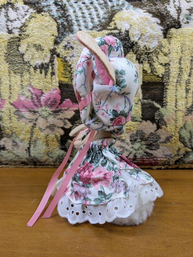 Puede incluir: Una figura de conejito de peluche que lleva un gorro y un vestido con estampado floral con detalles de cinta rosa. El vestido presenta un ribete de encaje de ojal blanco. El conejito tiene orejas largas y de color claro.