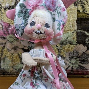 Puede incluir: Una mu&ntilde;eca de conejito hecha a mano con un gorro y un vestido florales. El gorro presenta rosas rosas y moradas sobre un fondo blanco. El vestido tiene un estampado floral a juego con un ribete de encaje de ojal. Una cinta rosa est&aacute; atada alrededor del cuello.