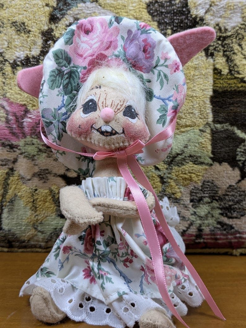 Puede incluir: Una mu&ntilde;eca de conejito hecha a mano con un gorro y un vestido florales. El gorro presenta rosas rosas y moradas sobre un fondo blanco. El vestido tiene un estampado floral a juego con un ribete de encaje de ojal. Una cinta rosa est&aacute; atada alrededor del cuello.