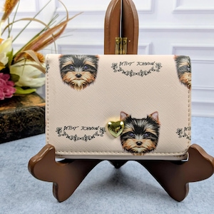 May include: A beige bi-fold wallet featuring a repeating print of Yorkshire Terrier faces and the text Betsey Johnson. The wallet is accented with a small, polished gold-tone heart-shaped stud centered on the front flap.