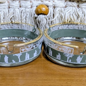 May include: Two clear glass ashtrays with a gold rim and a green band featuring white classical figures and trees. The ashtrays are round and sit on a wooden surface.
