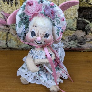 Puede incluir: Una mu&ntilde;eca de conejito hecha a mano con un gorro y un vestido florales. El gorro y el vestido tienen un fondo blanco con rosas rosas y moradas. La mu&ntilde;eca tiene una cara sonriente, orejas rosas y una cinta rosa.