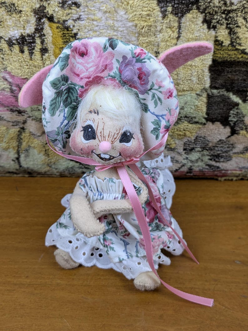 Puede incluir: Una mu&ntilde;eca de conejito hecha a mano con un gorro y un vestido florales. El gorro y el vestido tienen un fondo blanco con rosas rosas y moradas. La mu&ntilde;eca tiene una cara sonriente, orejas rosas y una cinta rosa.