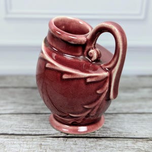 May include: A small, glossy, deep burgundy ceramic pitcher featuring a rounded body with an embossed leaf pattern. The vessel has a single curved handle attached to the side and a flared rim, finished with a smooth, reflective glaze throughout.