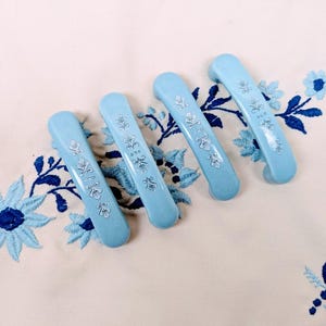 May include: Four light blue cabinet handles with a floral design. The handles are arranged on a cream-colored surface with blue floral embroidery. Each handle is approximately 5 inches long.