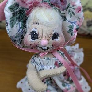 Puede incluir: Mu&ntilde;eca de conejito hecha a mano con un gorro y un vestido con estampado floral. El gorro presenta rosas rosas y moradas sobre un fondo blanco. La mu&ntilde;eca tiene una nariz rosa, ojos negros y una expresi&oacute;n sonriente. Una cinta rosa est&aacute; atada alrededor del cuello.