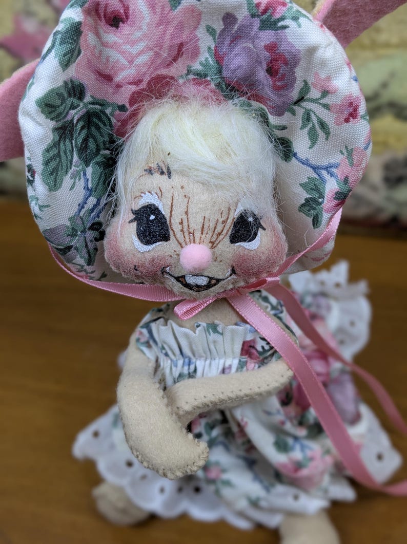 Puede incluir: Mu&ntilde;eca de conejito hecha a mano con un gorro y un vestido con estampado floral. El gorro presenta rosas rosas y moradas sobre un fondo blanco. La mu&ntilde;eca tiene una nariz rosa, ojos negros y una expresi&oacute;n sonriente. Una cinta rosa est&aacute; atada alrededor del cuello.